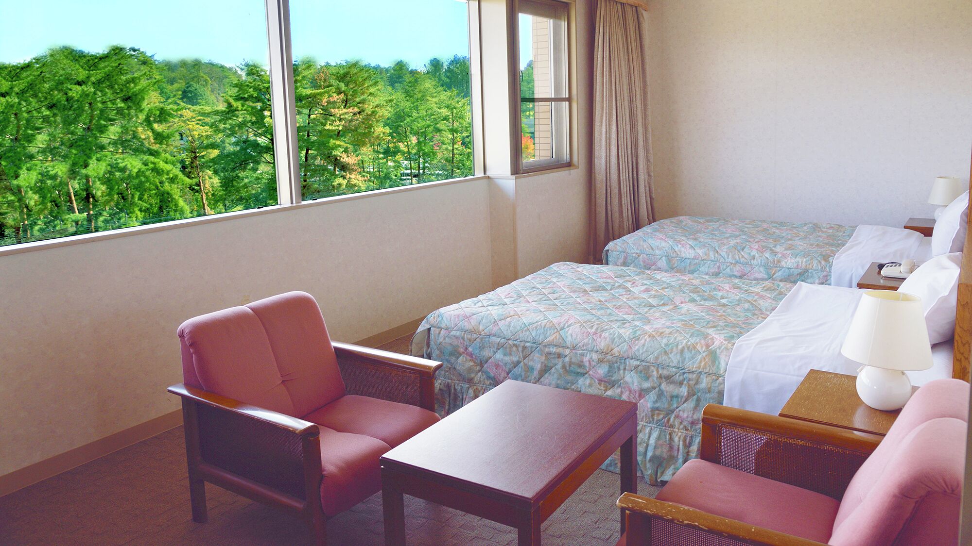 A hotel room with two beds, a chair, a table, and a view of a forest through the window.