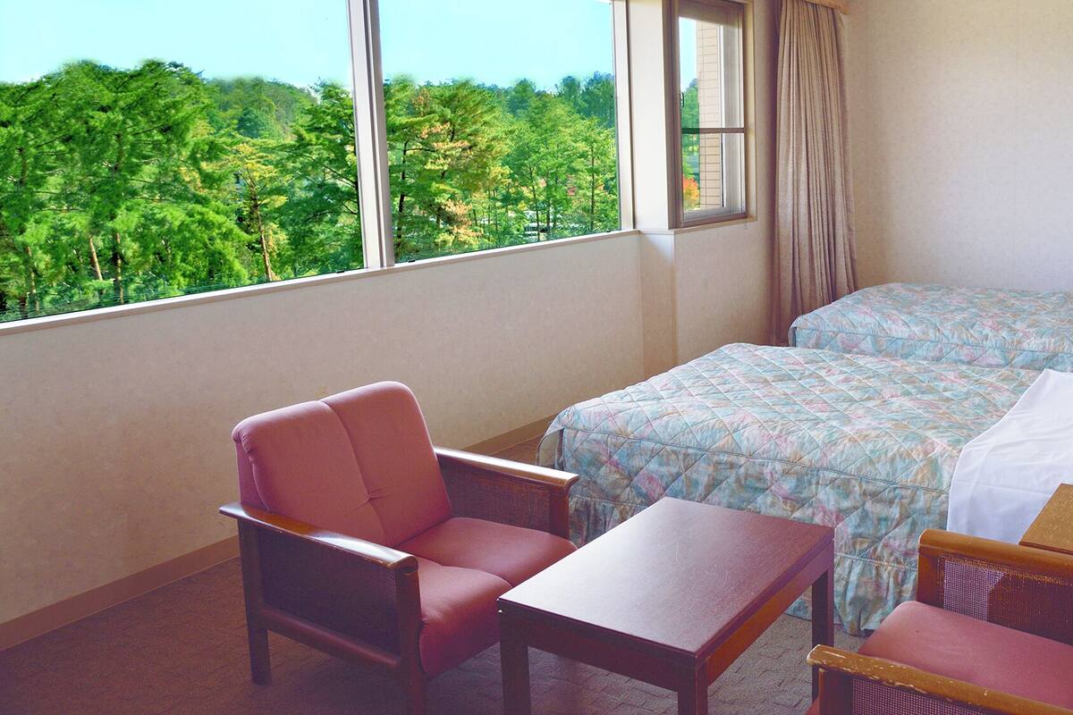 A hotel room with two beds, a chair, a table, and a view of a forest through the window.