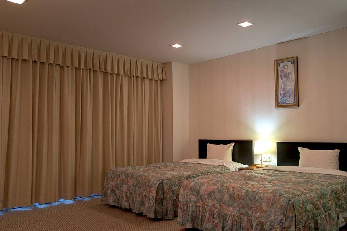 A hotel room with two beds, a large window with curtains, a painting on the wall, and a nightlight.