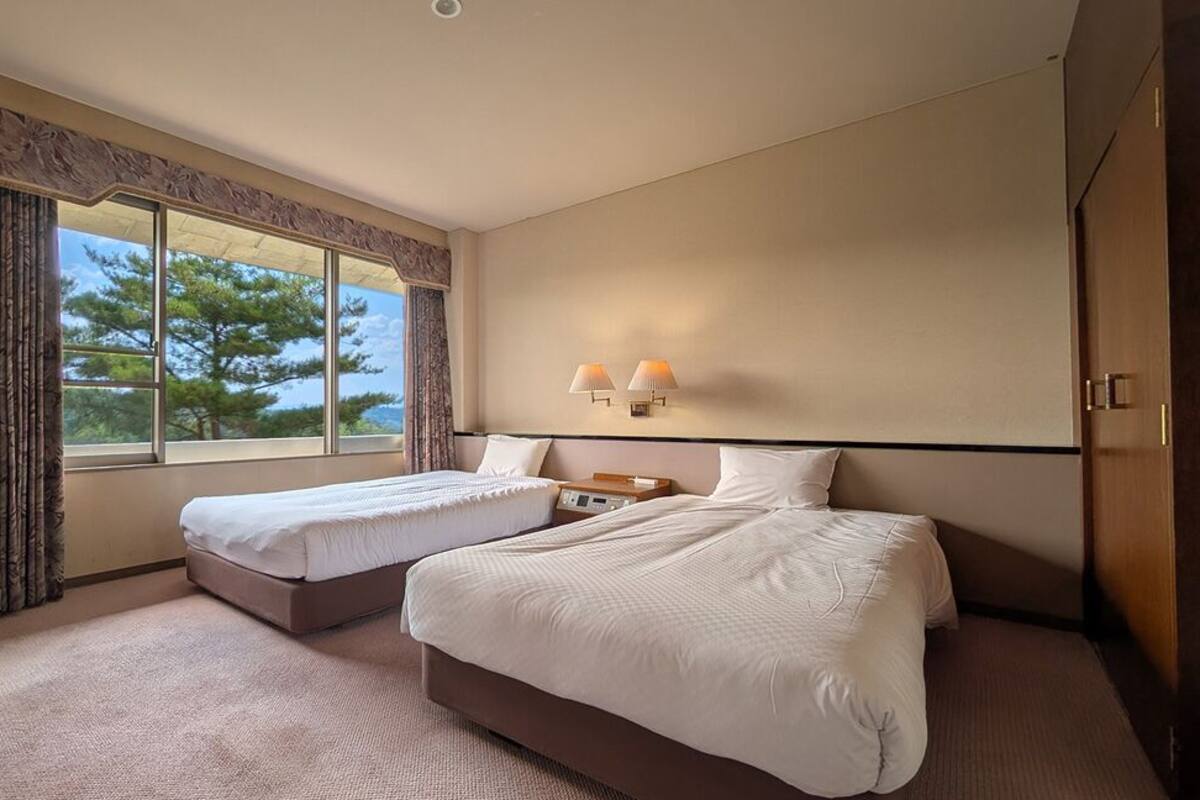 A hotel room with two beds, a TV, a desk, and a window with a view.