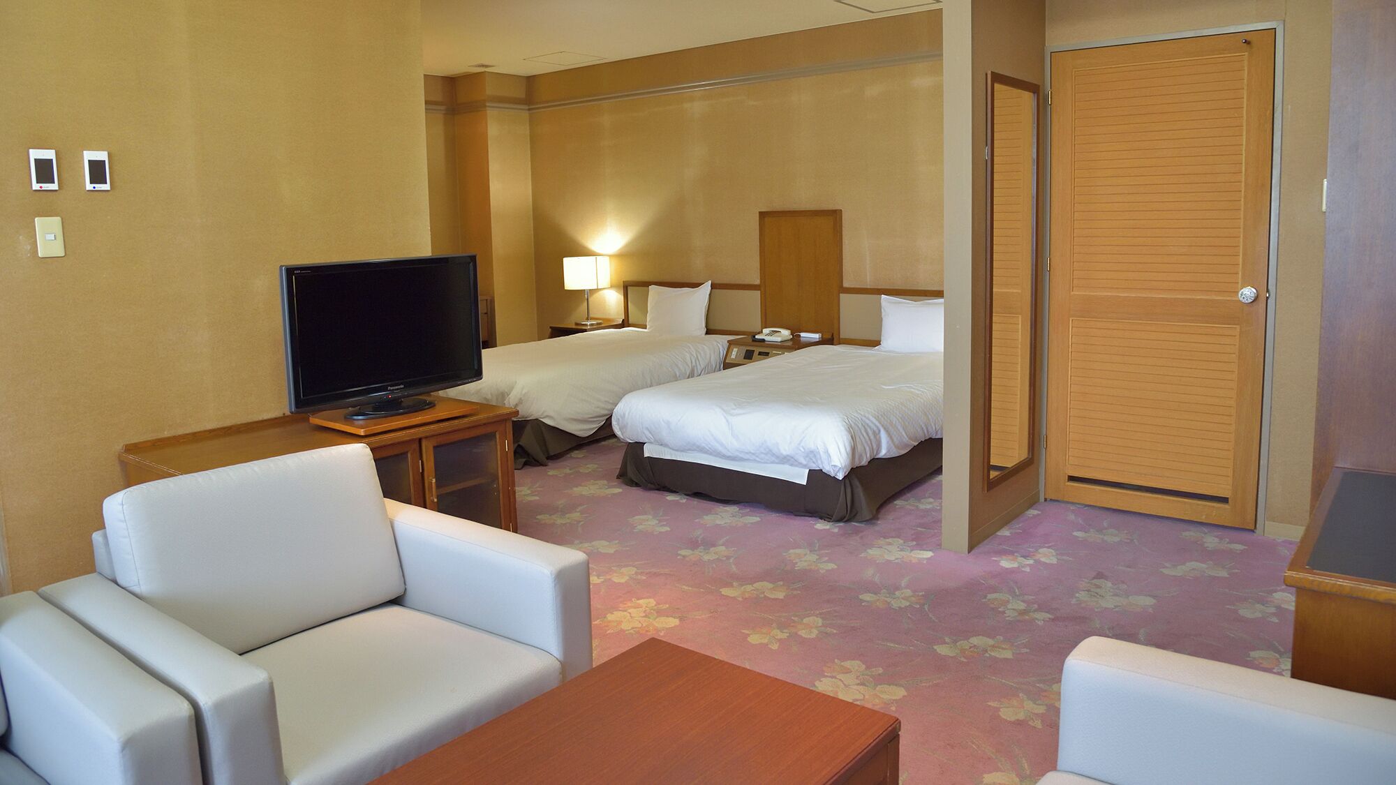 A hotel room with a television, two beds, a desk, and a seating area.