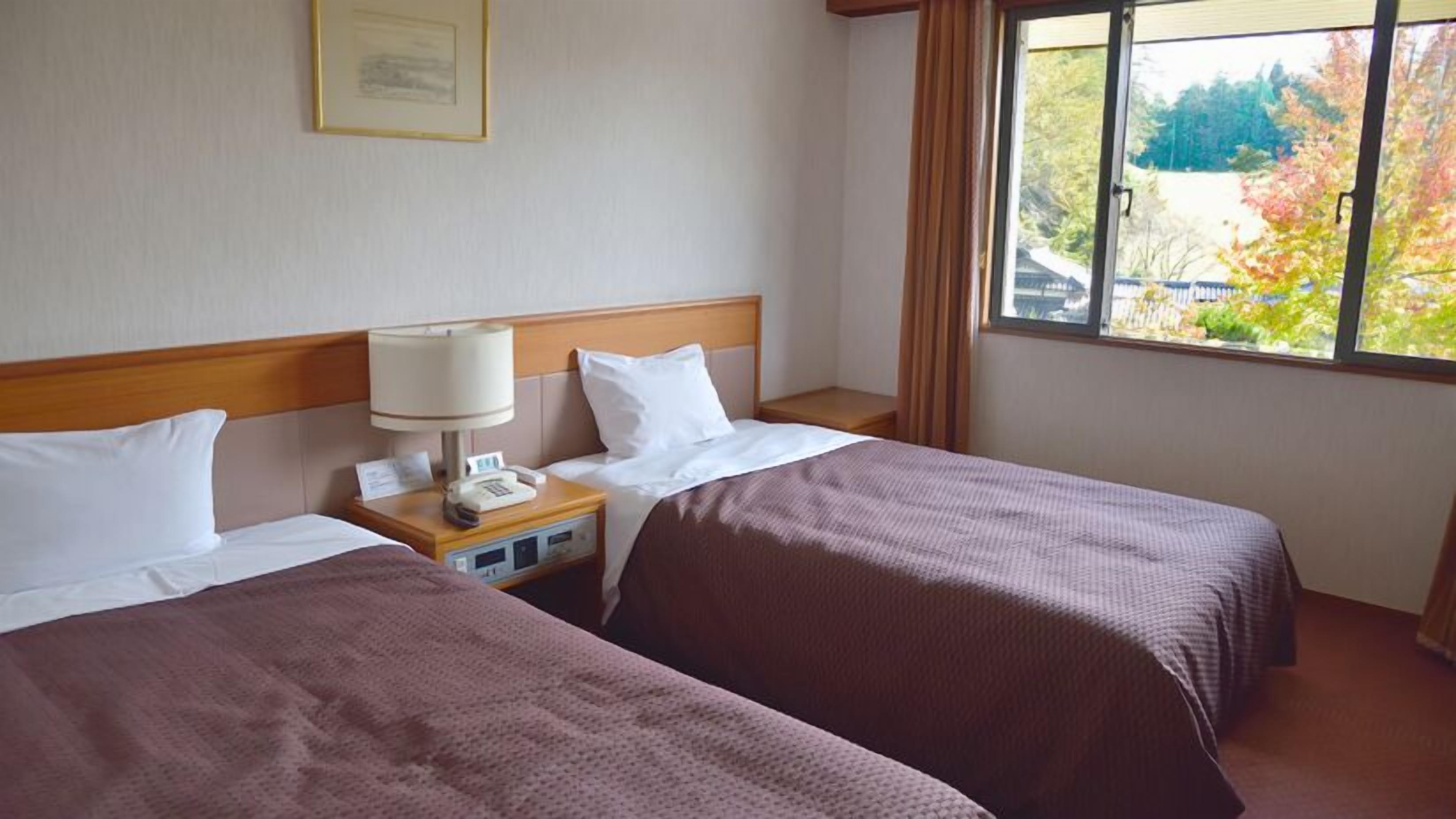 A hotel room with two beds, a nightstand with a lamp, a telephone, and a window with a view of trees.