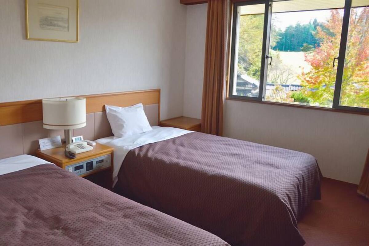 A hotel room with two beds, a nightstand with a lamp, a telephone, and a window with a view of trees.