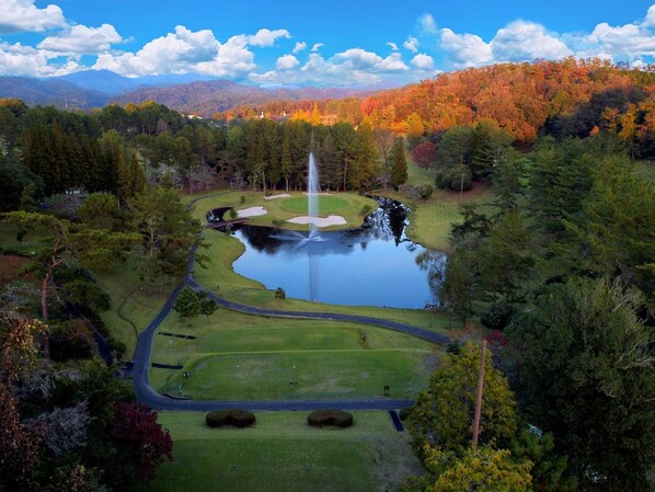 A golf course with a central pond, surrounded by trees and a mountain range in the background.