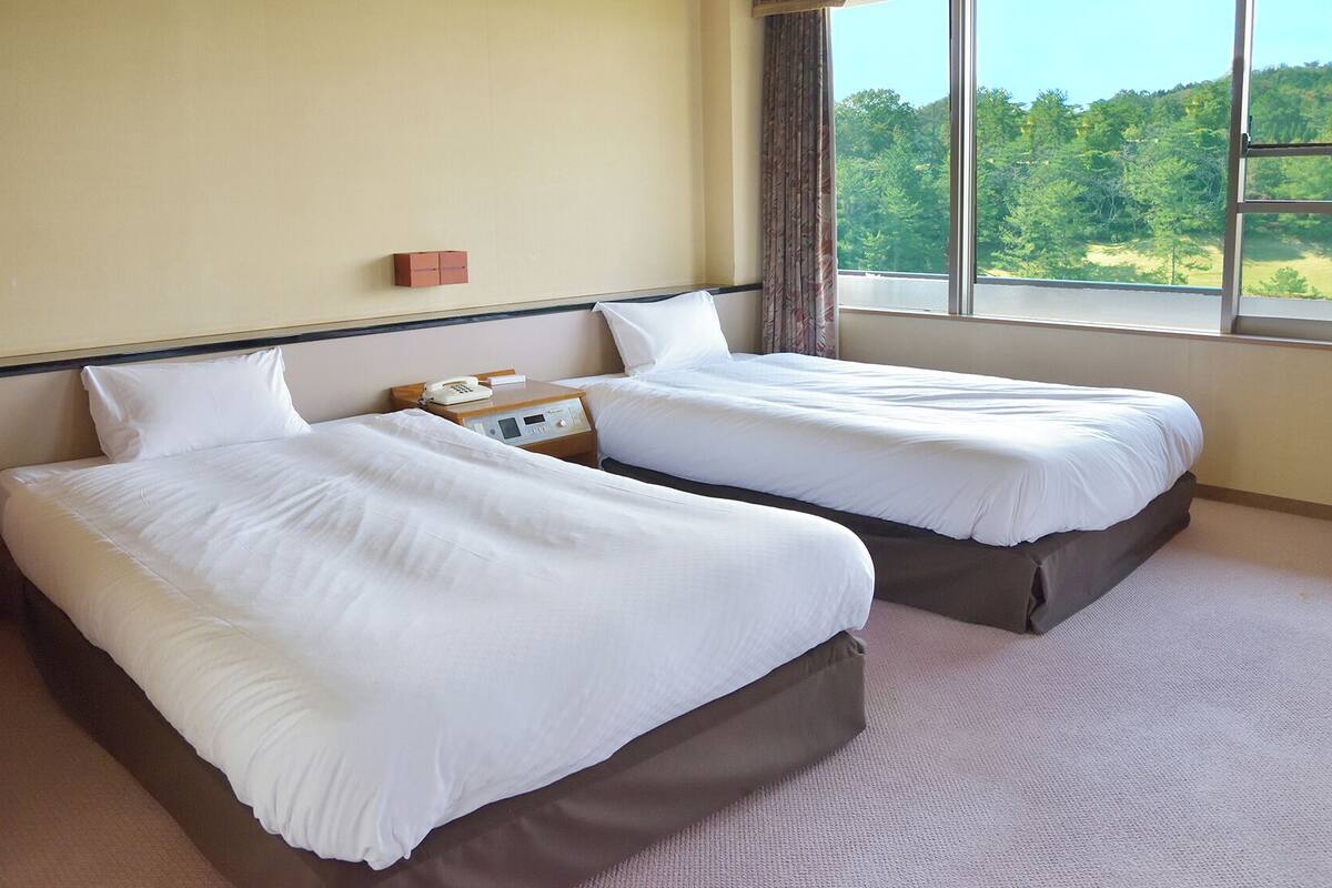 A hotel room with two beds, a bedside phone, and a window with a view of a forest.