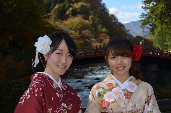 Two individuals in traditional attire standing in front of a bridge over a river, surrounded by autumn foliage. - Akari No Yado Villa Revage (Nikko)