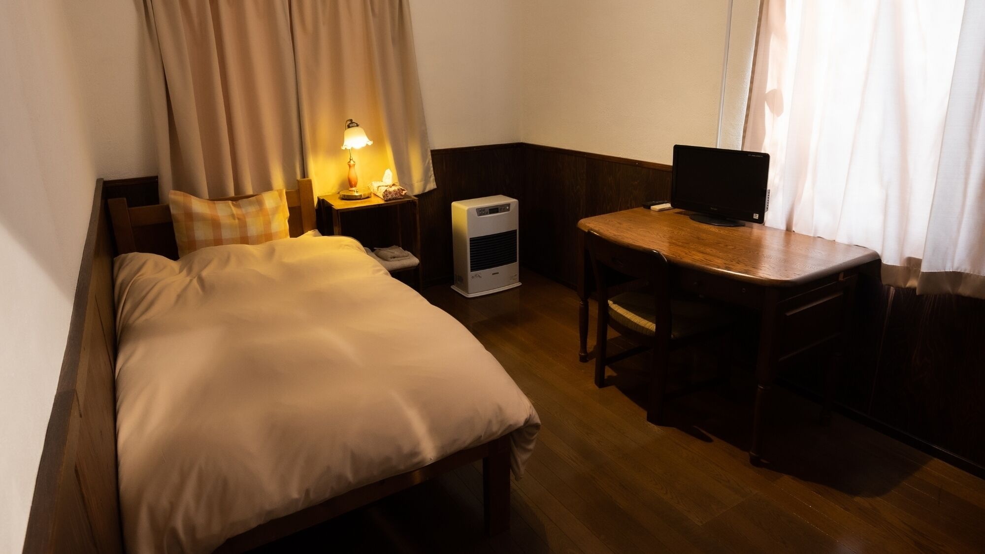 A hotel room with a bed, bedside table, lamp, desk with a computer, and a heater.