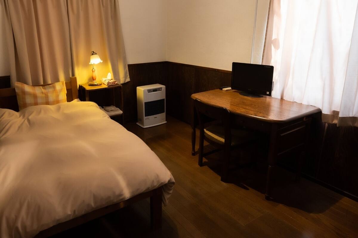 A hotel room with a bed, bedside table, lamp, desk with a computer, and a heater.
