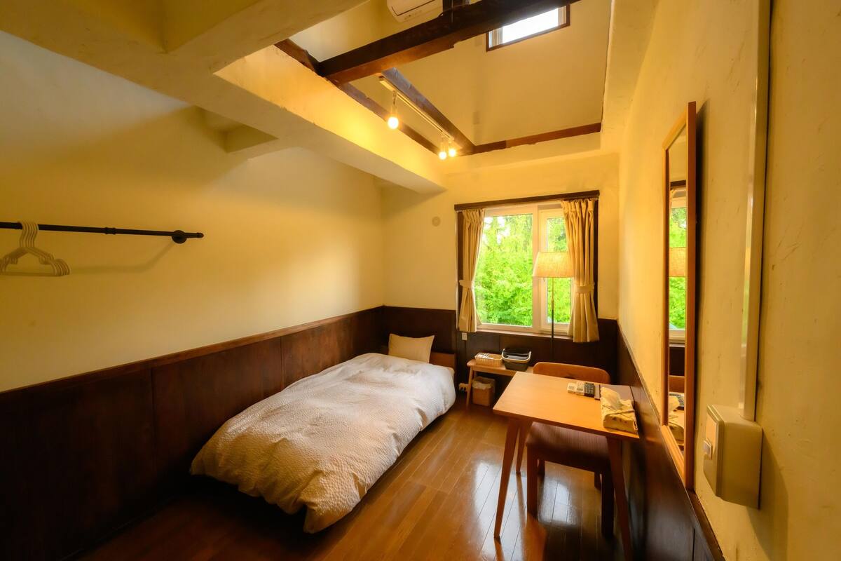 A room with a bed, a table, a chair, a window with curtains, and a hanging light fixture.