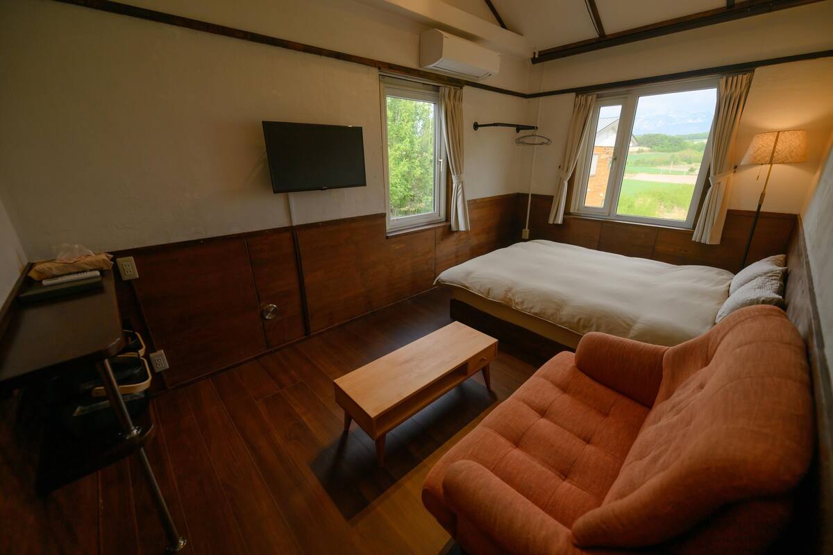 A room with a bed, a wooden bench, a TV mounted on the wall, and a window with curtains.