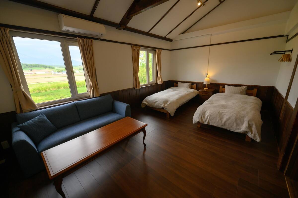 A room with a sofa, two beds, a wooden table, and a window with curtains.