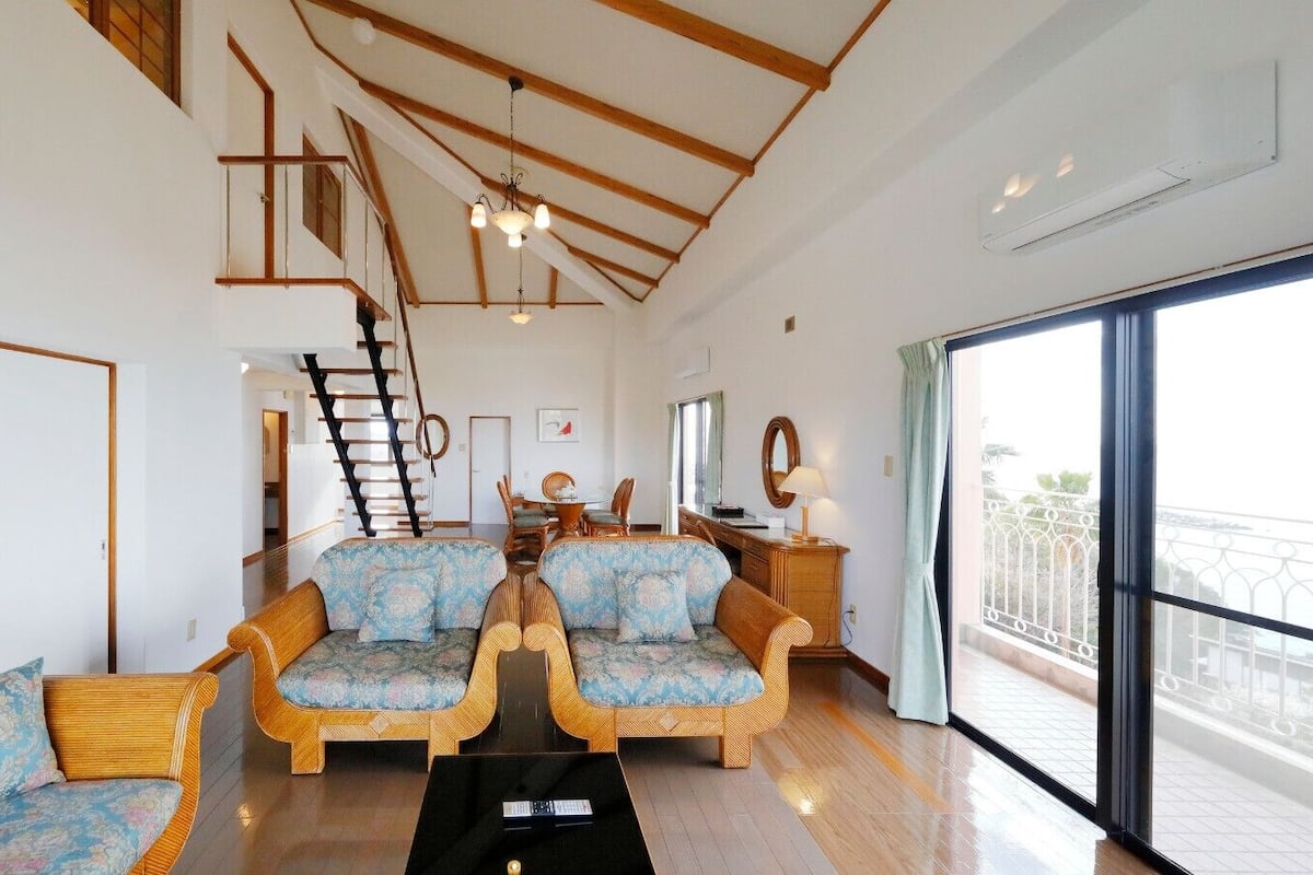 Amplia sala de estar con techo alto, vigas de madera, una escalera y un gran ventanal corredizo al exterior.