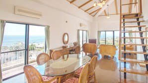 A spacious room with a large glass table surrounded by wicker chairs, a wooden staircase, and a view of the ocean.