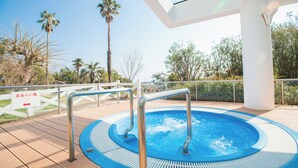 A hot tub on a wooden deck with metal railings, surrounded by palm trees and a white fence.
