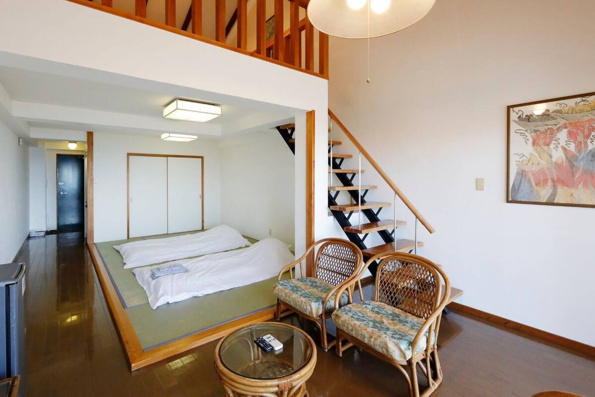 Una habitaci ón con una cama, dos sillas de mimbre, una mesita, un escritorio y una escalera.