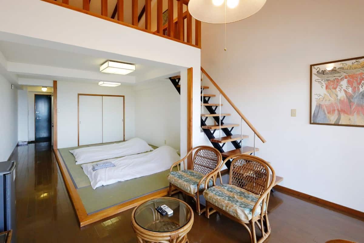 Una habitación con una cama, dos sillones de mimbre, una mesita, una escalera y un cuadro en la pared.