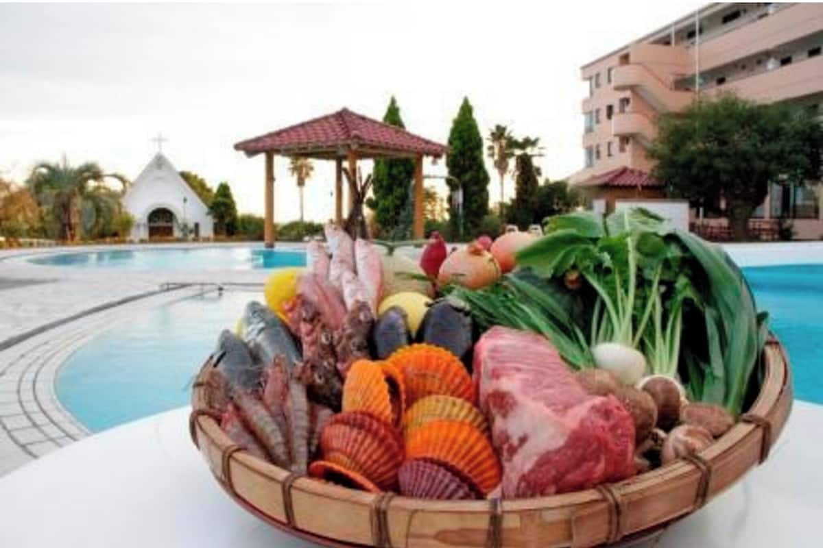 Zona de piscina con una cesta de mariscos, una pequeña iglesia blanca y un edificio de varios pisos al fondo.