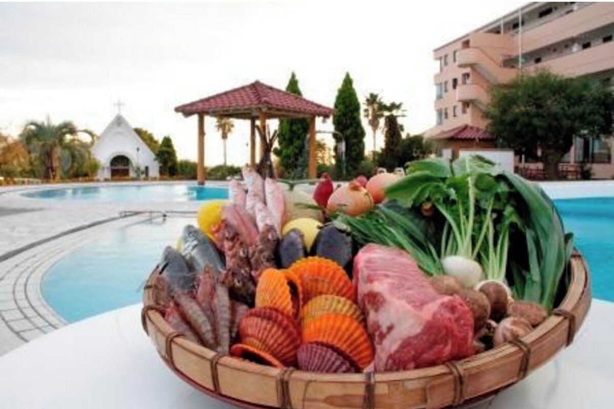 Un espace aménagé au bord de la piscine avec un panier rempli de fruits de mer, une petite église blanche et, au fond, un immeuble à plusieurs étages.
