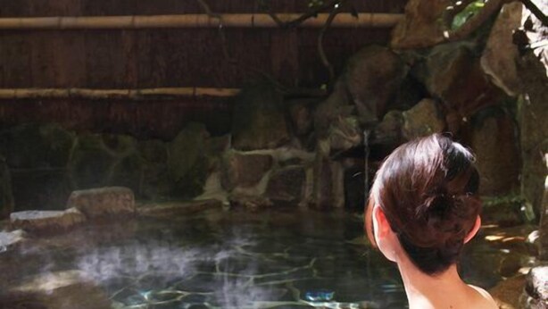 Una persona en un entorno de spa, con una bañera de piedra y una estructura de madera al fondo.