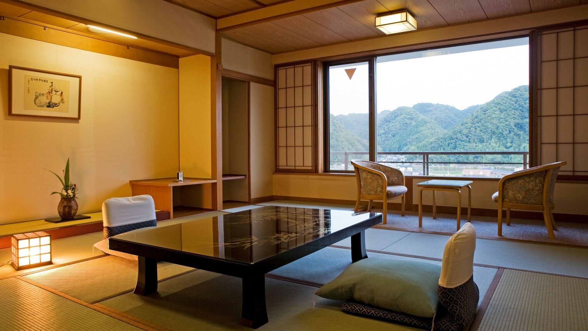 A traditional Japanese room with tatami flooring, a low table, chairs, a view of mountains, and a framed picture on the wall.