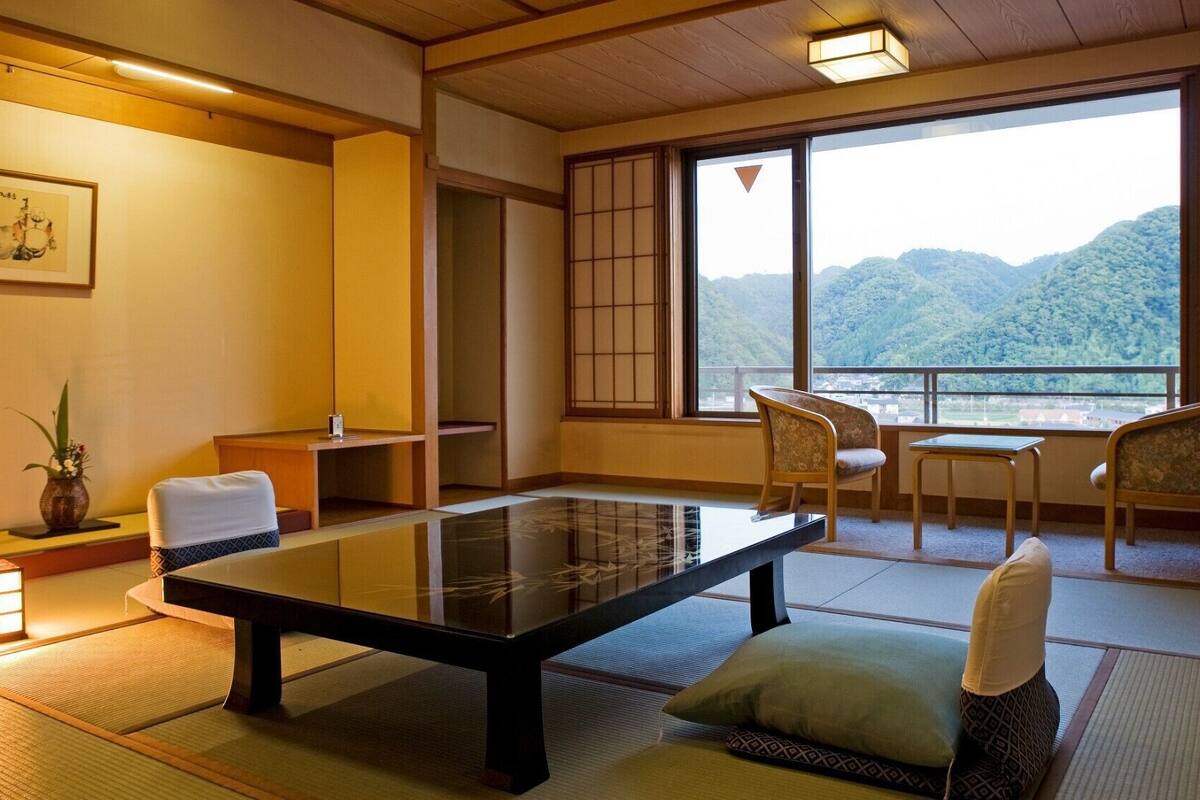 Um quarto japonês tradicional com tatames, uma mesa baixa, cadeiras, vista para montanhas e um quadro na parede.