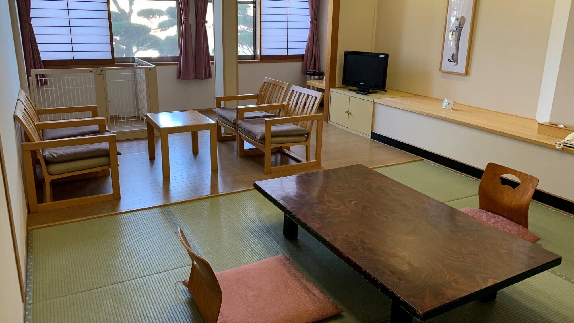 A traditional Japanese-style room with tatami flooring, a low table, wooden chairs, a television, and a window with a view of trees.