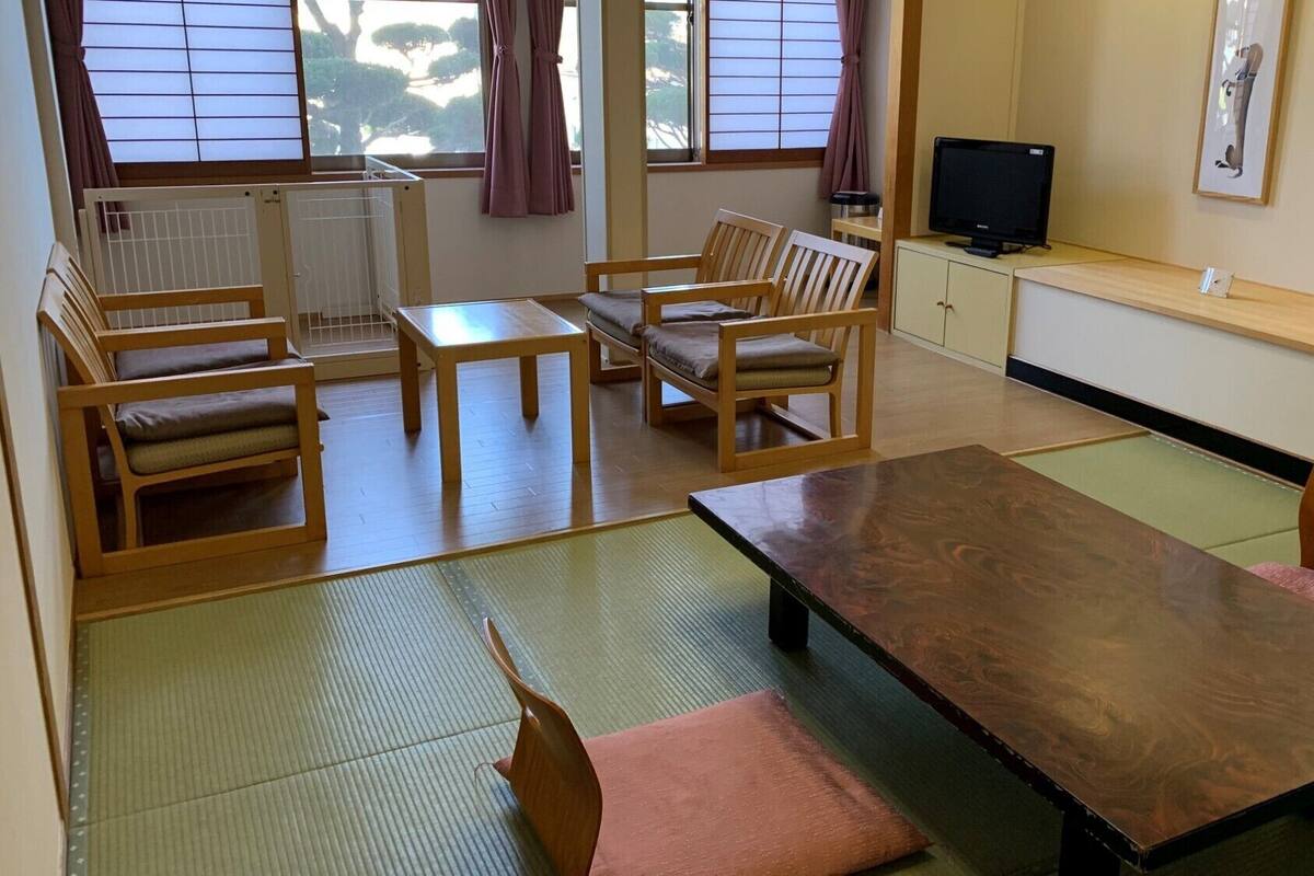 Um quarto tradicional japonês com tatames, uma mesa baixa, cadeiras de madeira, uma televisão e uma janela com vista para árvores.