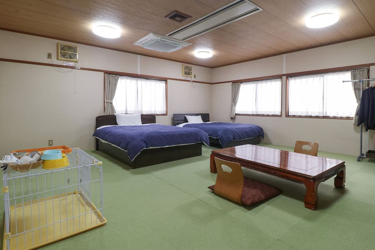 A room with two beds, a table, a chair, a small table with a basket, and a coat rack.