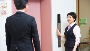 A man and a woman standing at an elevator, with a pink wall and a door handle visible. - Yakushido Spa (Oshu)