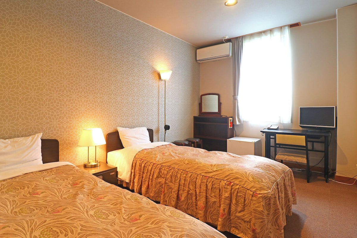 A hotel room with two beds, a desk, a chair, a TV, and a window with curtains.