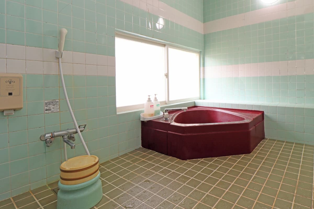 A bathroom with a bathtub, a toilet, a window, and a hand soap dispenser.