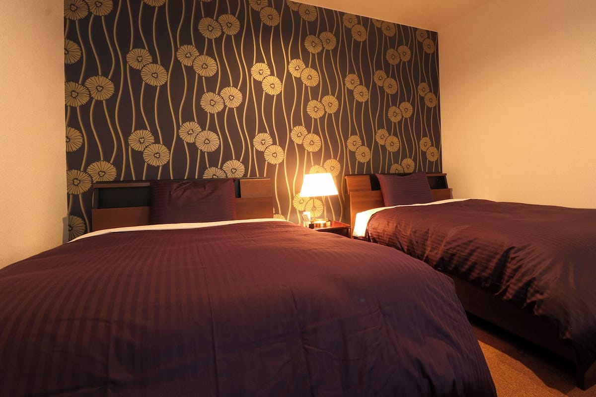 A hotel room with two beds, a nightstand with a lamp, and a floral wallpaper design.