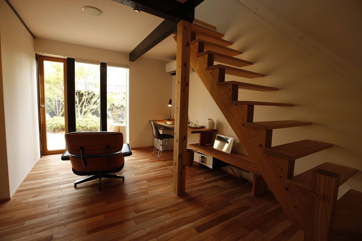 A spacious room with a wooden staircase, a desk with a chair, and a large window providing a view of the outdoors.