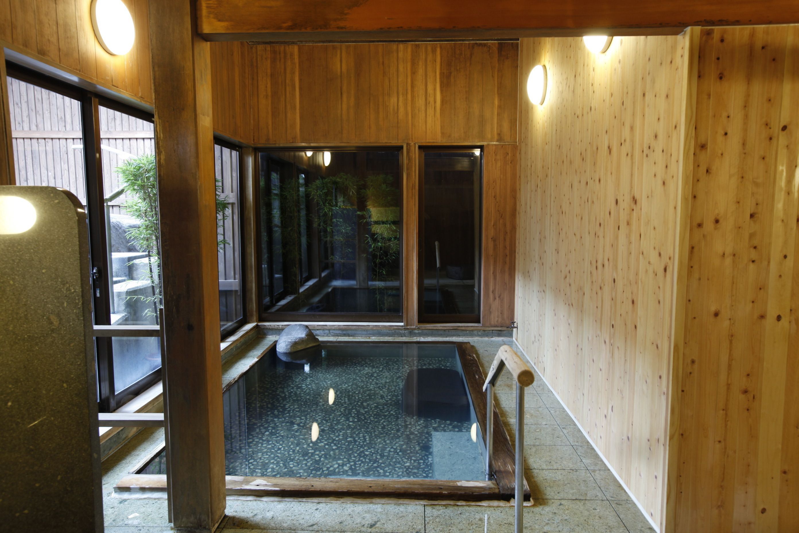 A traditional Japanese onsen with a wooden interior, a stone bath, and a view of the outdoors through large glass doors.