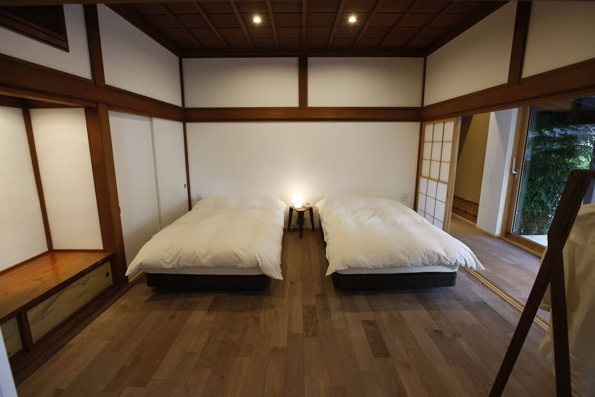 A traditional Japanese-style room with a wooden floor, two beds with white bedding, a small bedside table with a lamp, and a sliding door leading to a garden.