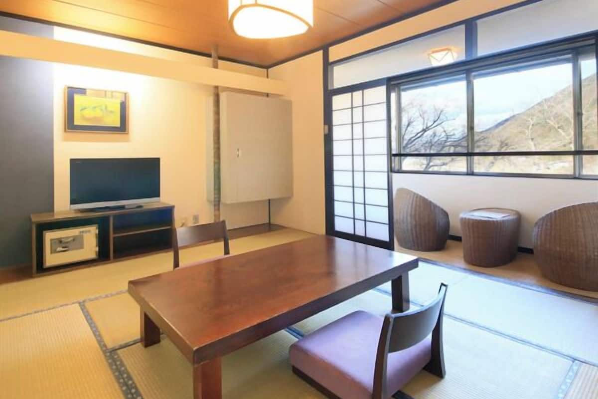 A traditional Japanese room with tatami flooring, a low table, a television, and a large window with a view of the outdoors.