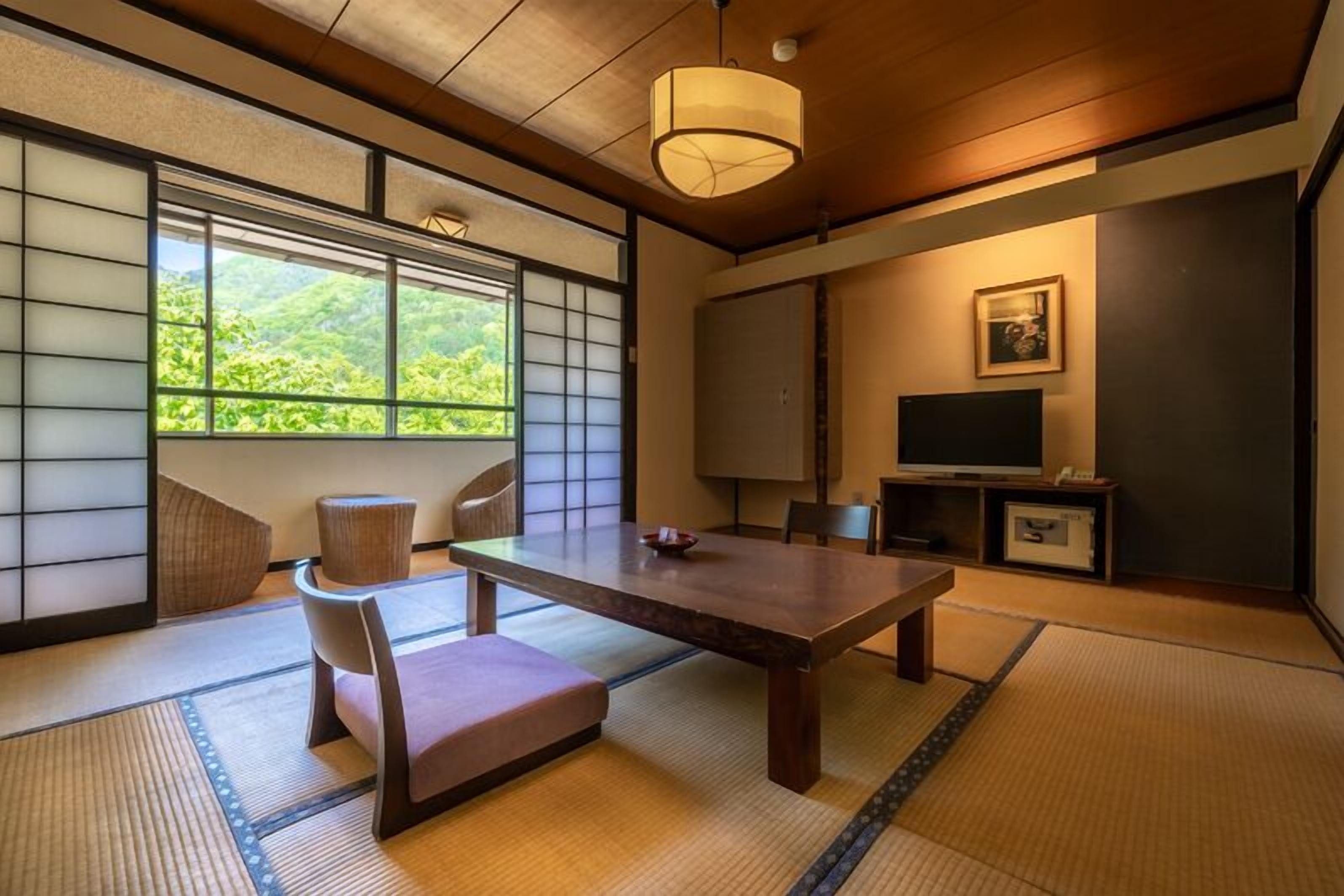 Une pièce de style japonais traditionnel, avec un sol recouvert de tatamis, une table basse, un fauteuil rembourré, un téléviseur et une porte coulissante donnant sur un paysage verdoyant.