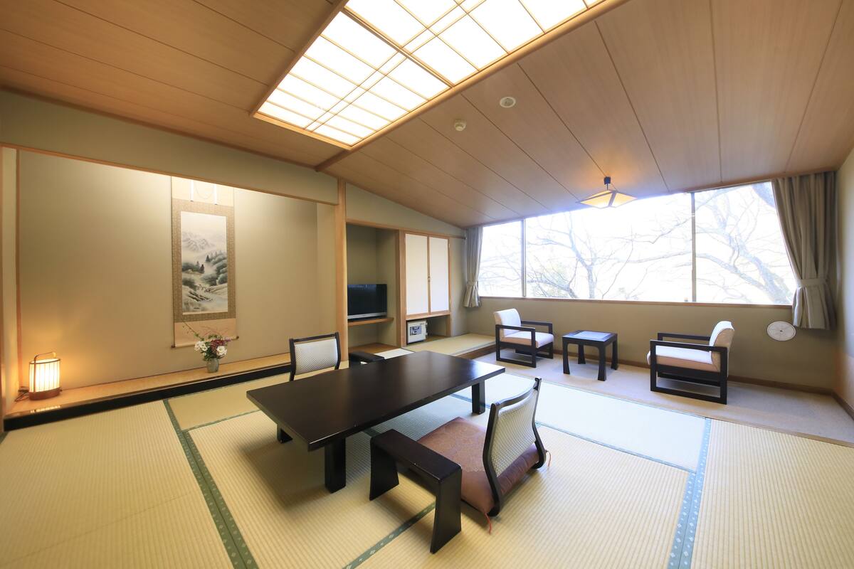 Una habitación tradicional japonesa con suelo de tatami, una mesa baja con cojines y un amplio ventanal con cortinas.