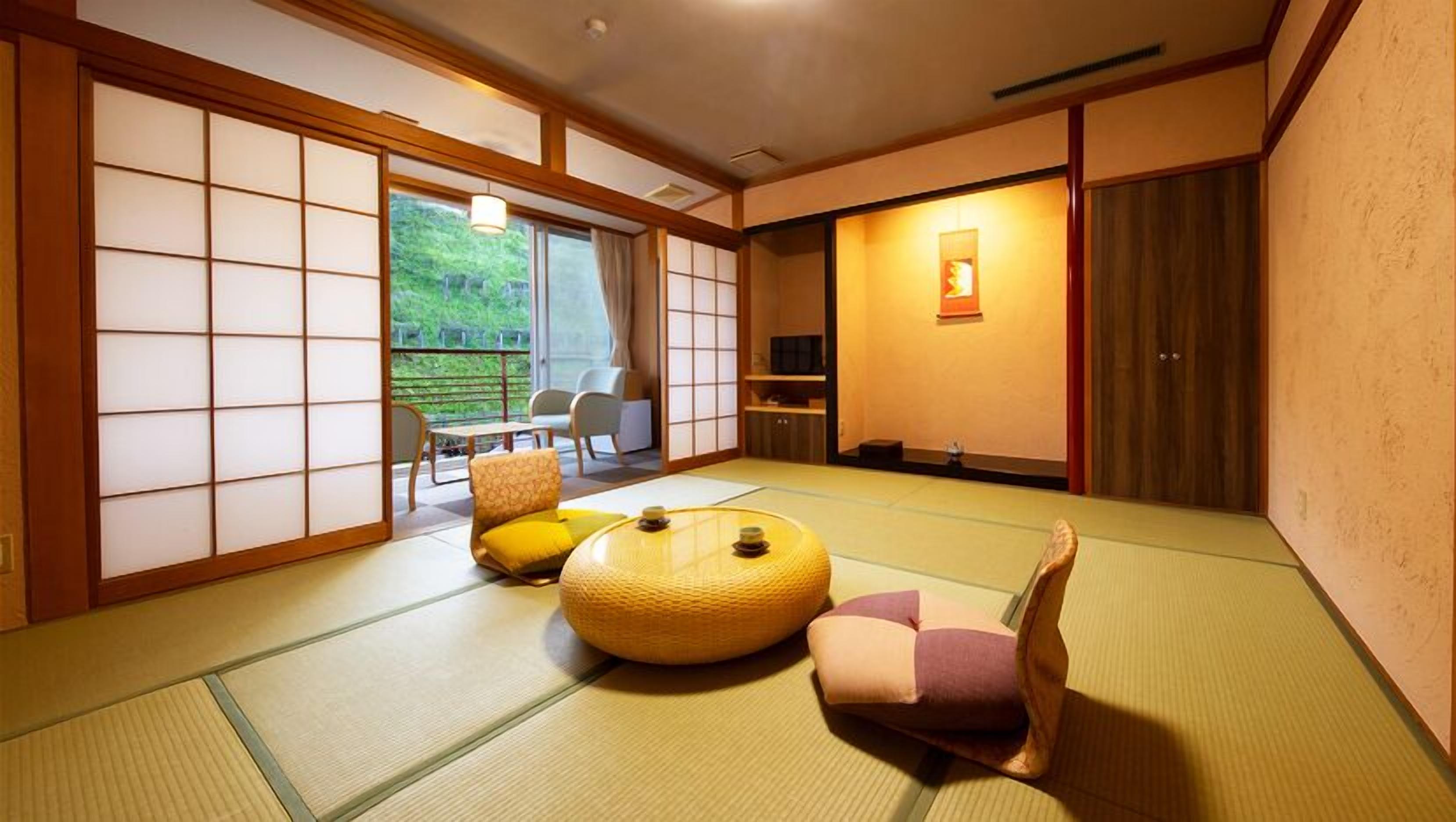 A traditional Japanese room with tatami flooring, a low table, cushions, sliding doors, and a view of greenery outside.