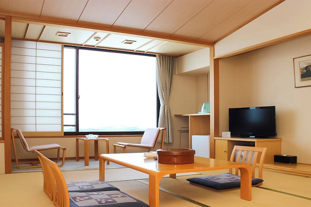 Una habitación tradicional japonesa con suelo de tatami, una mesa baja, sillas, un televisor y una ventana con vistas.