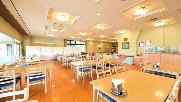 A hotel dining area with multiple tables and chairs, a whiteboard, and a kitchen area in the background.