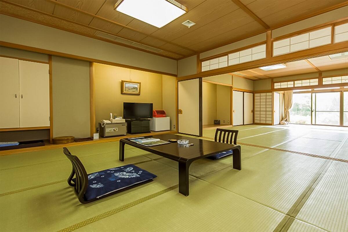 Una habitación japonesa tradicional con suelo de tatami, una mesa baja, sillas, televisor y puertas correderas.