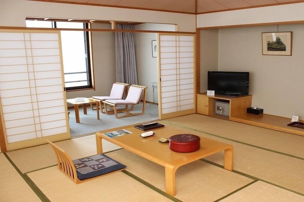 Una habitación tradicional japonesa con suelo de tatami, un televisor, una mesa pequeña con un recipiente rojo y una puerta corredera que da a una zona de estar.