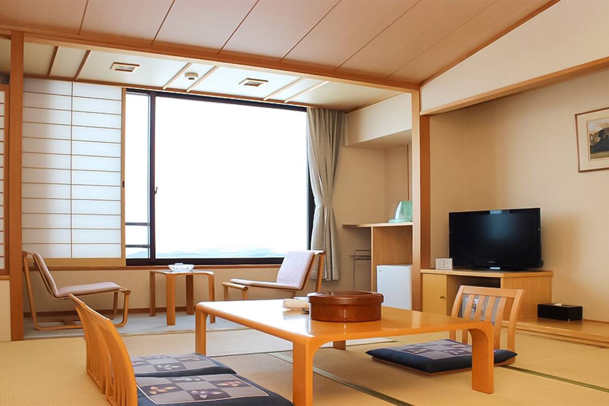 Una habitación tradicional japonesa con suelo de tatami, una mesa baja, sillas, un televisor y una ventana con vistas.