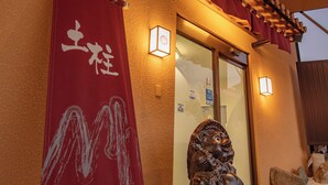 A building entrance with a red banner, a wooden pergola, and two wall-mounted lights. - Dochu Land Shin Onsen (Awa)