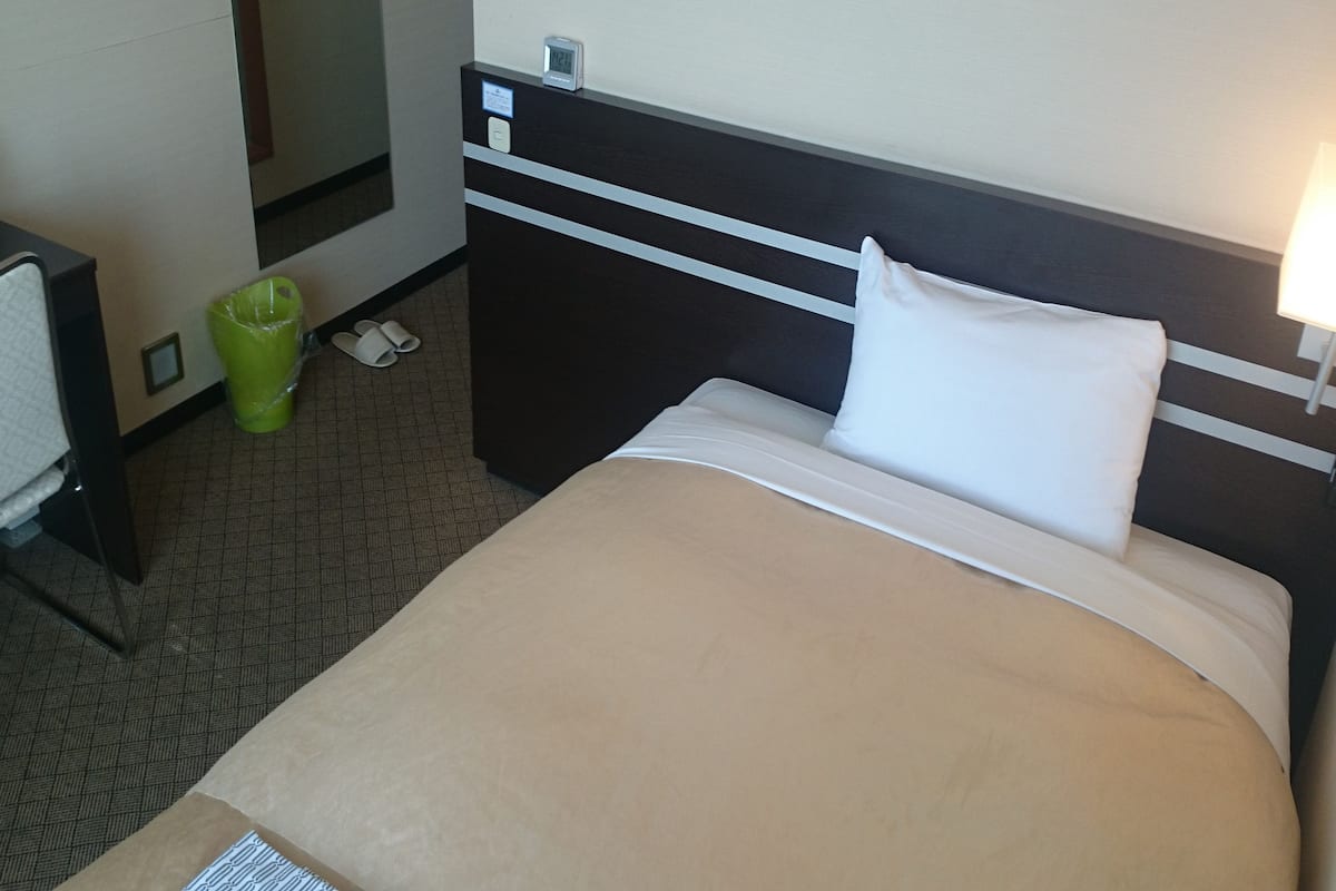 Een hotelkamer met een bed, nachtkastje, prullenbak, slippers en een wandklok.