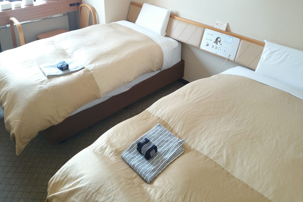 Twee enkele bedden met beige beddengoed, een houten hoofdbord, een klein bureau met stoel, en een bedieningspaneel aan de muur.