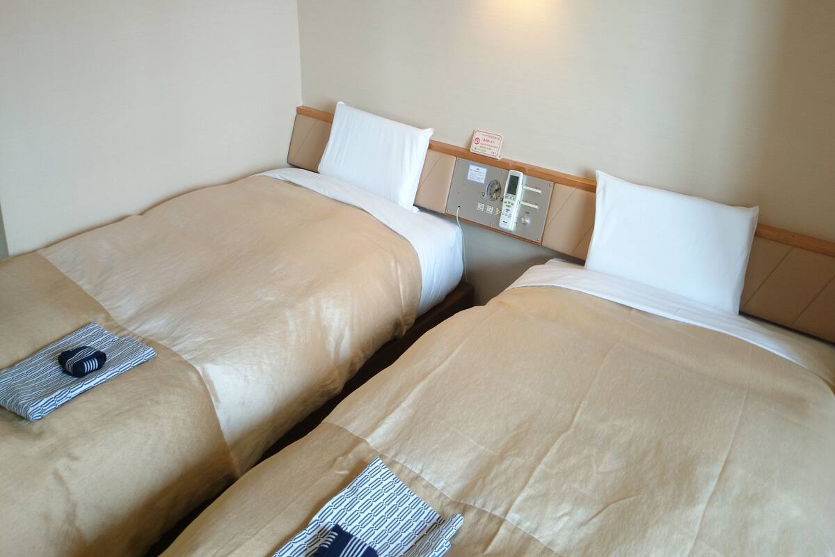 Twee enkele bedden met beige beddengoed, een nachtkastje met een lamp, en een bedieningspaneel aan de muur.