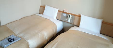 Two single beds with beige bedding, a bedside table with a lamp, and a control panel on the wall.