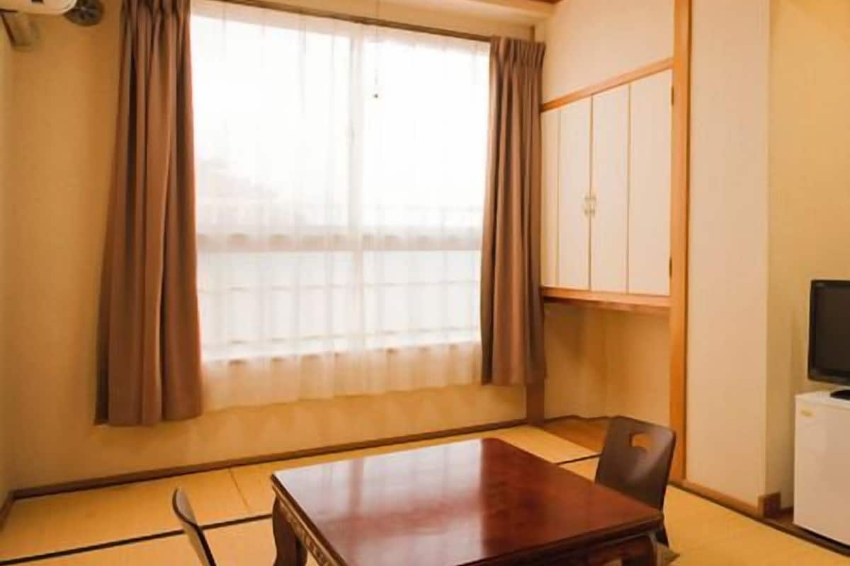 Ein Zimmer mit Tatami-Boden, einem Holztisch, einem Stuhl, einem Kleiderschrank und einem Fernseher.